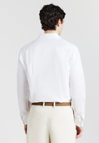 Uomo con capelli corti e ricci di colore scuro, che indossa una camicia bianca a maniche lunghe infilata in pantaloni beige chiaro con una cintura marrone, di spalle.