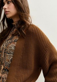 Brown knitted cardigan with a textured pattern, featuring wide sleeves and a loose fit. Paired with a patterned blouse underneath.