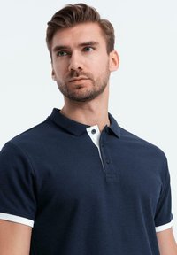 Marineblauw polo met witte kraag en mouwaccenten, voorzien van een drieknoopsluiting en een gestructureerde stofafwerking.