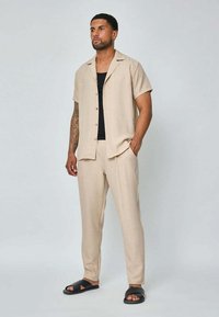 Chemise col beige clair à manches courtes, avec un devant boutonné, associée à un pantalon assorti. Tissu texturé et sandales à glissière noires.