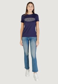 T-shirt blu navy con logo "GUESS" in dettagli brillanti, abbinato a jeans denim azzurri e sneakers bianche. Tessuto morbido e design aderente.