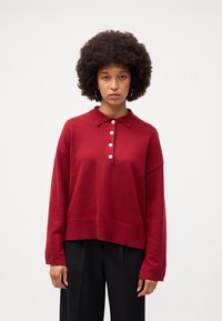 Pulôver polo tricotado vermelho com pala de botões, ombros caídos e bainha canelada. Textura suave e ajuste descontraído, combinado com calças pretas.