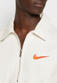 Veste blanche à fermeture éclair avec col, présentant un logo Nike orange brodé sur le côté gauche et une texture lisse et mate.