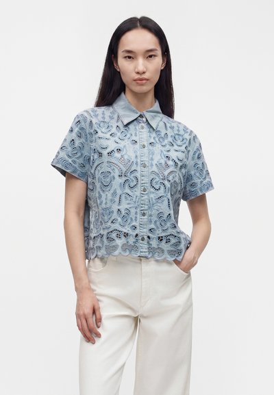 Jeune femme aux longs cheveux foncés porte une chemise bleue brodée à manches courtes boutonnée et un pantalon blanc, avec une main dans la poche.