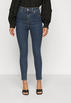 Kobieta ubrana w niebieskie obcisłe jeansy z wysokim stanem, czarną bluzkę z długim rękawem oraz czarne sandały na obcasie z odkrytymi palcami, stojąca na tle jednolitego, jasnego tła.