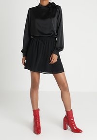 Abito nero con top in satin e vita elastica, gonna fino al ginocchio. Abbinato a stivaletti rossi lucidi con tacco a blocco.