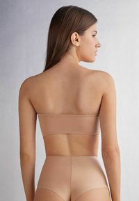 Nude bandeau-top en hoog-gesneden onderbroek van een gladde, rekbare stof. Voorzien van subtiele stiksel-details. Achteraanzicht toont het minimalistische ontwerp.