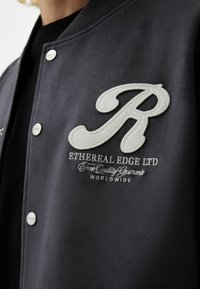 Black varsity jacket featuring a textured, oversized white "R" patch, embroidered brand name and details, and snap button closure.