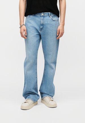 Person trägt hellblaue Jeans mit geradem Bein, schwarzes Oberteil, weiße Turnschuhe und ein silbernes Armband am linken Handgelenk, steht vor einem einfarbigen Hintergrund.