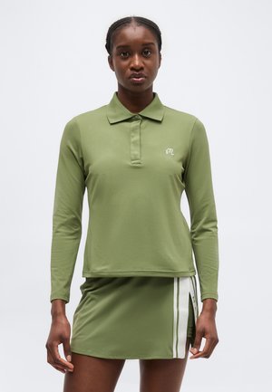Donna che indossa una polo a maniche lunghe verde oliva e una gonna abbinata con strisce bianche laterali, in piedi davanti a uno sfondo semplice.