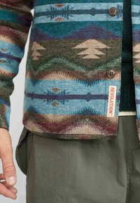 Multikleuren geweven jas met turquoise, bruin en crème patronen; heeft knopen en een "REVOLUTION" label aan de zoom.
