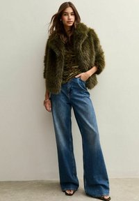 Młoda kobieta w oliwkowej sztucznej futrzanej kurtce, w camouflagedowej bluzce, szerszych niebieskich dżinsach i czarnych sandałach na obcasie, stojąca na tle jednolitej ściany.