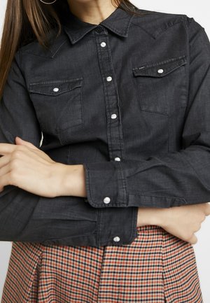 Femme portant une chemise gris foncé boutonnée avec deux poches poitrine et une jupe à carreaux, croisant les bras sur son corps.