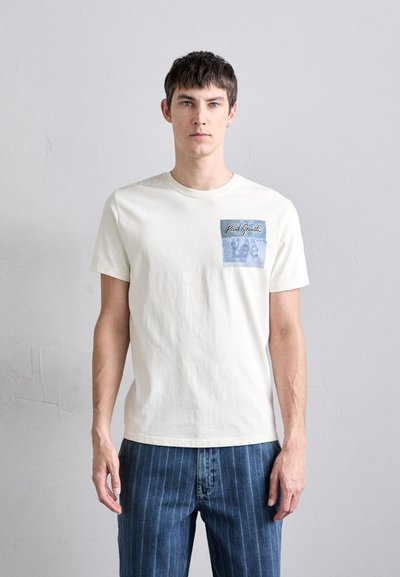 Tricou din bumbac de culoare crem, cu guler rotund și mâneci scurte, având un patch grafic albastru pe partea stângă a pieptului.