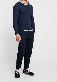 Maglione strutturato blu navy sopra una camicia colletto azzurro chiaro, abbinato a pantaloni scuri e sneakers nere con dettagli bianchi e logo.