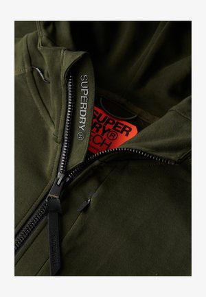 oliwkowy hoodie z pełnym zamkiem błyskawicznym, z logo "SUPERDRY" na uchwycie zamka i kołnierzu, z czerwonym metkiem wewnątrz dekoltu.