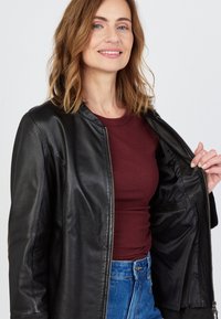 Musta nahkatakki, jossa on vetoketju edessä, pyöreä kaula-aukko ja teksturoitu pinta. Päällä burgundinpunainen yläosa ja yhdistettynä sinisiin farkkuihin.