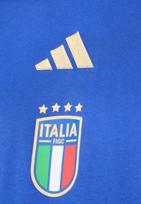 Tricou sportiv albastru cu logo-ul Adidas brodat în aur și emblema echipei naționale de fotbal a Italiei cu patru stele deasupra.