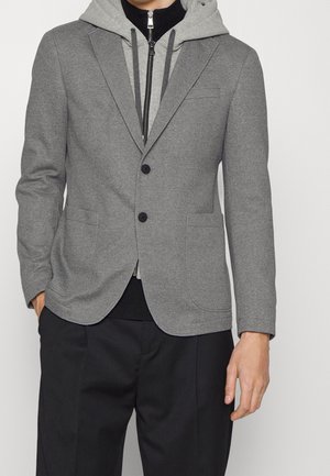 Hombre vistiendo un blazer gris sobre una sudadera con capucha gris con cremallera y pantalones negros, con una mano en el bolsillo, contra un fondo blanco.