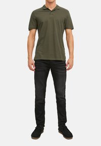 Olivgrünes Polohemd mit Kragen, kurzen Ärmeln und gesticktem Logo, kombiniert mit dunklen Jeans und schwarzen Schnürschuhen.