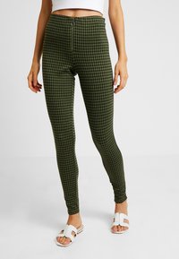 Gröna rutiga leggings med hög midja och frontdragkedja, med en åtsittande design och slät textur. I kombination med vita sandaler.