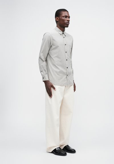 Stripete skjorte i beige og mørkebrun, kombinert med kremfargede vide bukser og svarte loafers, på en modell mot en lys bakgrunn.