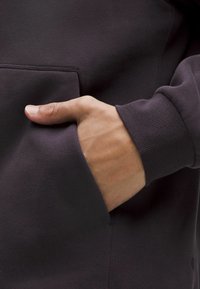 Sötét kapucnis pulóver, puha tapintással, oldalzsebbel és bordás mandzsettákkal. A zsebben pihenő kéz a funkciót és illeszkedést szemlélteti.