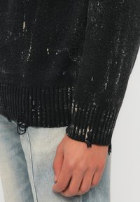 Fekete kötött pulóver fehér, kopott mintákkal. Bordázott mandzsetták és szegélyek, rojtos széllel. Világoskék farmernadrág fölött viselve.