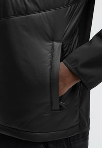 Chaqueta negra con textura acolchada, bolsillo con cremallera en el lado izquierdo y un diseño ajustado con mangas elegantes.