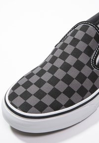Chaussure slip-on à motifs damiers noirs et gris avec une tige en toile, une semelle en caoutchouc blanche, et des détails de couture subtile le long des bords.