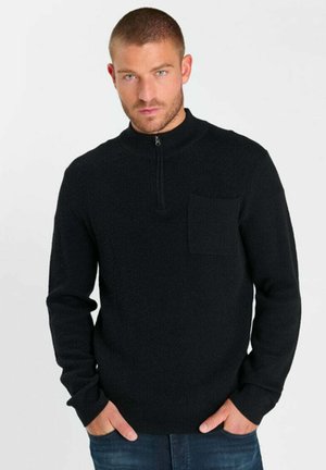 Uomo con i capelli corti che indossa un maglione nero a maniche lunghe con tasca sul petto e mezza zip, in piedi con le mani nelle tasche.