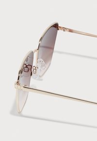 Goldfarbene Flieger-Sonnenbrille mit grauenbraunen Gläsern, ausgestattet mit Metallrahmen, transparenten Nasenpolstern und schmalen Bügeln.