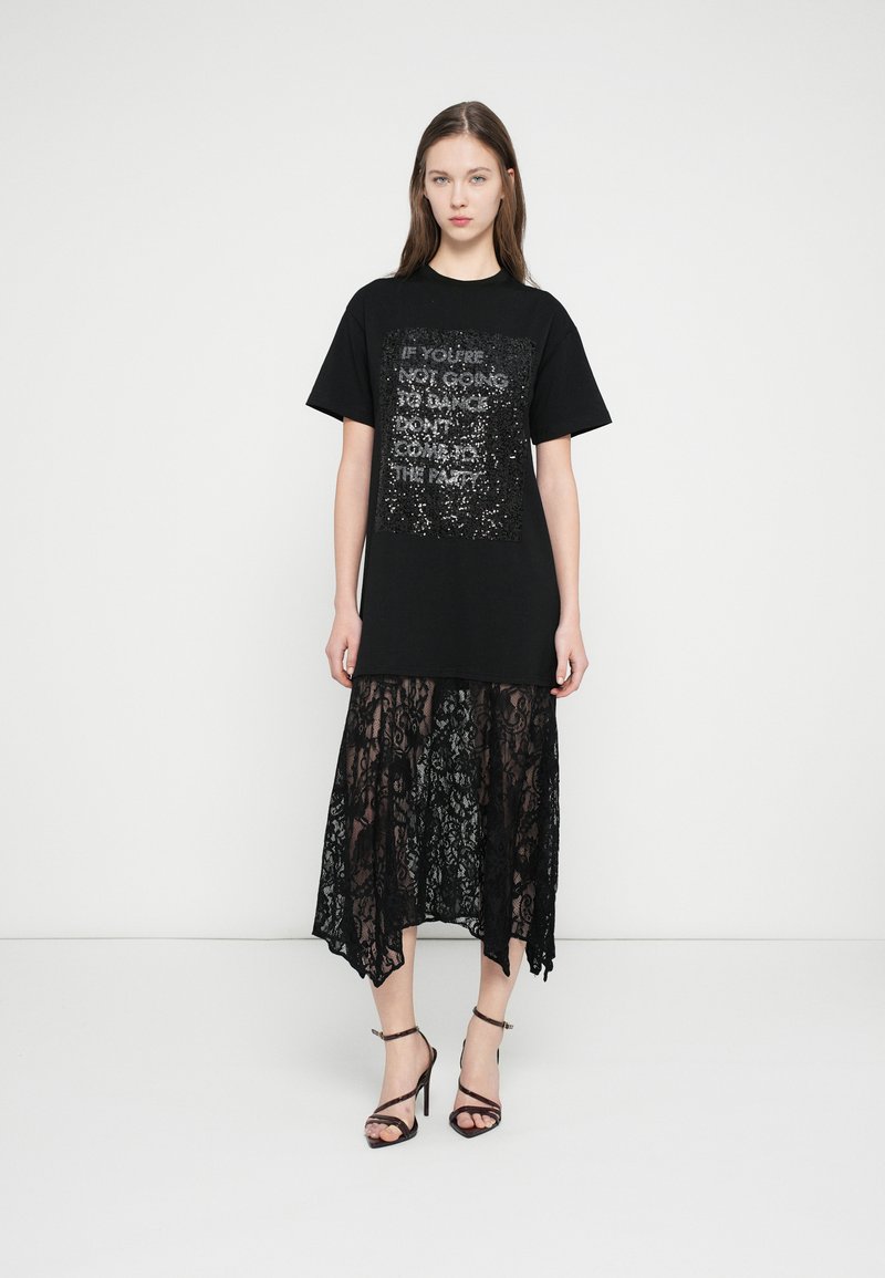 Juoda oversize stiliaus marškinėliai su sidabrinių blizgučių grafika, dėvimi virš permatomos juodos nėriniuotos suknelės su banguotais kraštais. Juodi bateliai ant aukštos platformos su petnešėlėmis.