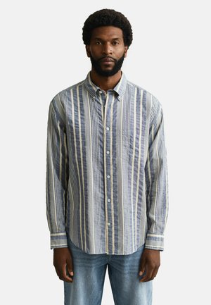 Mann steht frontseitig, trägt ein blau-weiß gestreiftes, längs gestreiftes Hemd mit Knopfleiste und hellblaue Jeans vor einem schlichten Hintergrund.