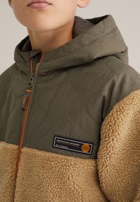 Pihezüst zöld dzseki barna polár betéttel, elöl cipzárral, narancssárga díszítéssel és fekete címkével, fehér felirattal.