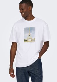 Άσπρο βαμβακερό T-shirt με εκτυπωμένη εικόνα του Πύργου του Άιφελ σε απαλούς χρωματισμούς. Λαιμόκοψη τύπου crew και κοντομάνικο, συνδυασμένο με σκούρο τζιν.