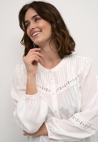 White blouse featuring a button-down front, vertical stripes, lace accents, and puffed sleeves with elastic cuffs. Soft, lightweight fabric.