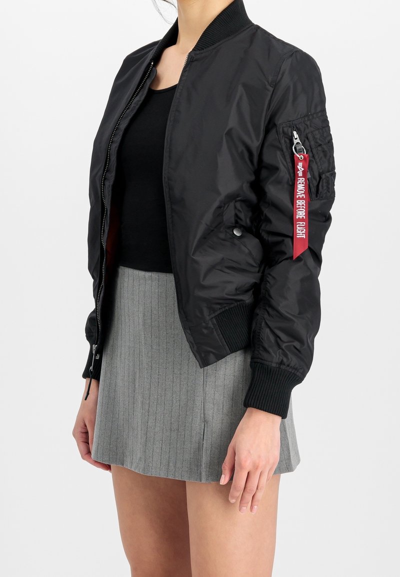 Femme portant une veste bomber noire avec une étiquette rouge "Remove Before Flight" sur la manche, un haut noir et une jupe grise côtelée sur fond blanc.