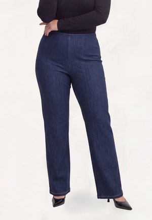 Kvinde iført mørkeblå jeans med lige ben, sort langærmet top og sorte spidse højhælede sko, der står med armene over kors.