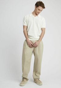 Polo shirt court en beige clair texturé à manches courtes associé à un pantalon crème ample et des chaussures beiges claires, se tenant devant un fond neutre.