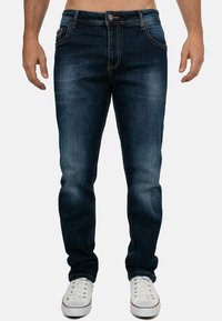 Jeans in denim blu scuro con vestibilità slim, caratterizzati da leggere sfumature, dettagli di cucitura arancione e design a cinque tasche. Indossati con sneakers bianche.