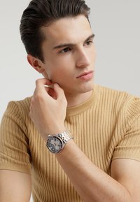 Silver- och roséguldarmband med en rund svart urtavla, metallband och kronograffunktioner, bärs på ett handled med en beige ribbad skjorta.