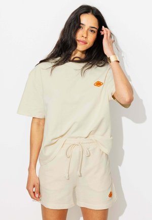 Femme aux longs cheveux foncés portant un t-shirt beige oversize et un short assorti avec cordon de serrage, touchant ses cheveux d'une main, fond neutre.