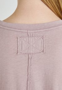 Svijetloružičasta t-shirt s pogledom sa stražnje strane, koja ima ušiveni pravokutni etiketu s X uzorkom i središnjim šavom.