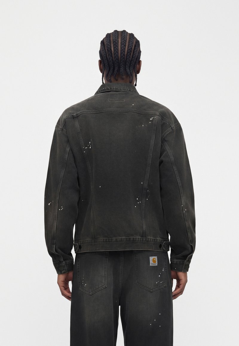 Man with braided hair wearing a black distressed denim jacket and matching pants, standing facing away against a plain background.