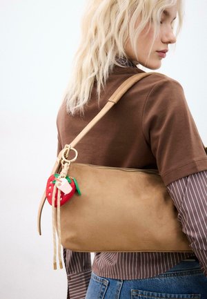 Donna bionda con maglie marroni a strati e jeans che porta una borsa a tracolla beige con un portachiavi a forma di fragola rossa.