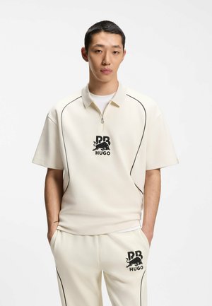 Junger Mann trägt ein cremefarbenes kurzärmeliges Polo mit Reißverschluss und passende Hosen mit schwarzem RB HUGO-Logo und Seitennähten, Hände in den Taschen.