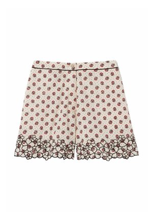 Mujer joven con top marrón sin mangas, pantalones cortos con estampado floral y borde festoneado, zapatos marrones, sosteniendo un bolso crema con detalle de lazo.