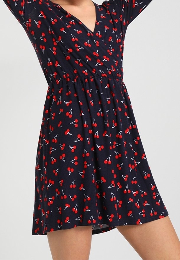 Femme portant une robe noire avec des motifs de cerises rouges et un col en V, les bras levés au-dessus de la tête, montrant une longueur mi-cuisse.