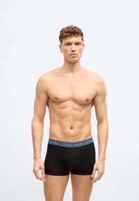 Άνδρας χωρίς πουκάμισο με σγουρά μαλλιά, φορώντας μαύρα εσώρουχα Tommy Hilfiger, στέκεται μπροστά σε απλό φωτεινό φόντο.